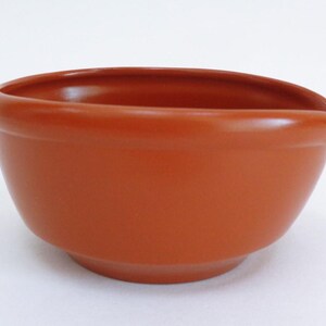 Puede incluir: Un cuenco de cerámica color terracota con forma redondeada y un pequeño pico en un lado. El cuenco tiene un acabado liso y brillante y se asienta sobre una superficie blanca. Este artículo de cocina es adecuado para servir o mezclar.
