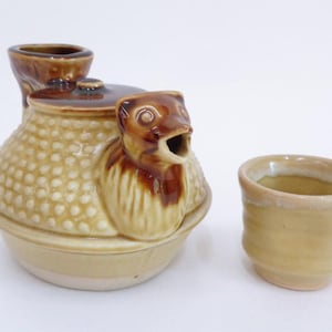 Puede incluir: Un juego de sake de cerámica con un servidor de sake marrón y beige con un pico en forma de pájaro y una taza a juego. El servidor tiene un cuerpo texturizado y una tapa pequeña. La taza tiene un diseño simple y redondeado.