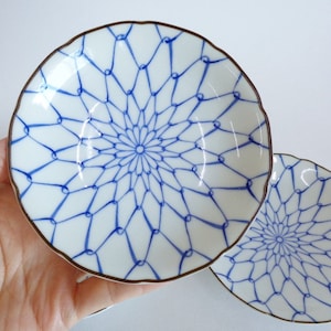 May include: Two white ceramic dishes with a blue geometric pattern and a brown rim. The dishes have a scalloped edge. One dish is held in a hand, and the other is in the background.