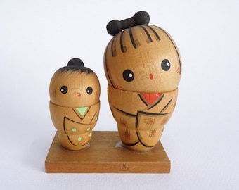 May include: Two wooden Japanese kokeshi dolls, one larger than the other, with painted faces and traditional clothing. The dolls are standing on a wooden base.