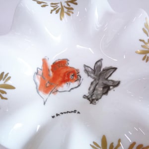 Puede incluir: Un plato de cerámica blanca con borde festoneado, decorado con pan de oro e imágenes pintadas a mano de dos peces. El plato está firmado "Katunisa".
