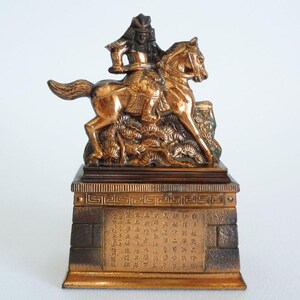 May include: A bronze-toned metal figurine depicting a samurai warrior on horseback, mounted on a rectangular base. The base features a central panel with engraved Japanese characters and a textured brick-like pattern on the sides.