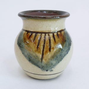 May include: A small, round ceramic vase with a cream-colored base and a dark brown interior. The exterior features a decorative pattern of brown and blue brushstrokes. The rim is dark brown, and the vase has a simple, elegant design.
