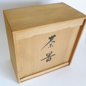May include: A rectangular light brown wooden storage box with a sliding lid. The front panel features black Japanese calligraphy characters printed vertically in the center. The box is constructed from natural wood with visible grain patterns.