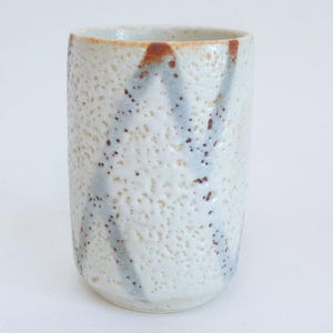 May include: A handmade ceramic tumbler with a textured, off-white surface. The tumbler features streaks of light blue and brown accents. The cylindrical shape is ideal for beverages. The pottery has a rustic, artisanal aesthetic.