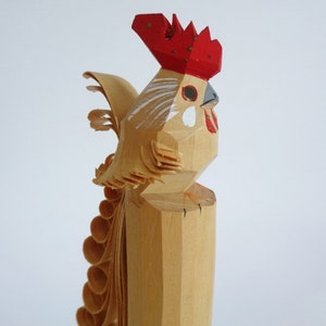 Könnte beinhalten: Eine hölzerne Hahnfigur mit rotem Kamm und Kehllappen, weißen Maldetails und einem geschnitzten Schwanz. Der Hahn steht auf einem Holzsockel.