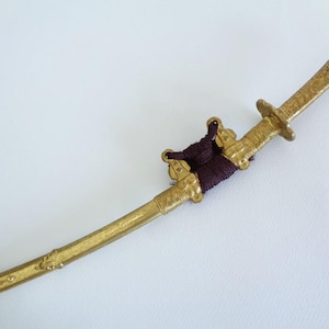 May include: A gold-colored metal sword scabbard featuring intricate engraved patterns along its length. The scabbard includes a dark purple cord wrapped around the center section, secured by two metal fittings, and a small circular guard near the handle.