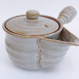 Puede incluir: Una tetera de cerámica de color beige claro con tapa y boquilla incorporada. La tetera tiene un asa acanalada y un pequeño pomo en la tapa. La boquilla es curva e inclinada.