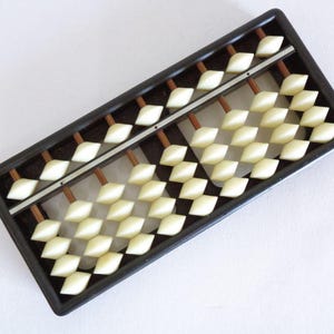 May include: A black abacus with rows of ivory-coloured beads. The beads are diamond-shaped and arranged on wooden rods within a rectangular frame. The abacus is a traditional calculating tool.