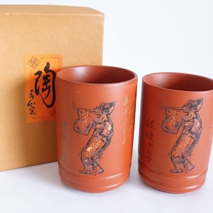 May include: Two terracotta-colored ceramic cups with a black illustration of a person playing a flute. The cups have Japanese text and are next to a cardboard box with a label featuring Japanese characters.
