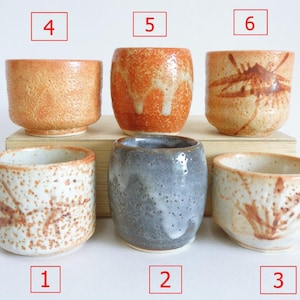 May include: Six ceramic cups with different glaze patterns. The cups are arranged in two rows of three. The cups are all about 7.5 cm tall and 5 cm wide. The cups are all made of a light brown clay. The glaze patterns are all different and include a speckled pattern, a brown and white pattern, a gray pattern, and a brown and orange pattern.