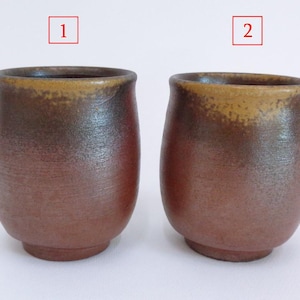 May include: Two handmade ceramic cups with a rustic, earthy aesthetic. The cups have a rounded shape with a slightly flared rim. The exterior features a gradient of brown and reddish-brown hues, with a lighter, golden-brown rim.