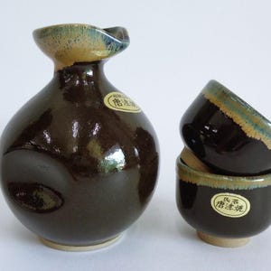 Puede incluir: Un juego de tres tazas de sake de cerámica marrón y una botella de sake. Las tazas y la botella tienen un esmalte marrón oscuro con un borde marrón claro y azul. La botella tiene una etiqueta con caracteres japoneses.