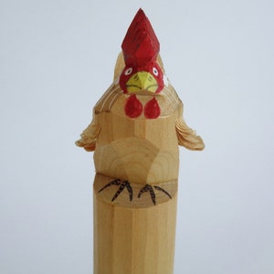 Könnte beinhalten: Eine hölzerne Hahnfigur mit rotem Kamm und Kehllappen, gelbem Schnabel und schwarz bemalten Füßen. Der Hahn steht aufrecht auf einem zylindrischen Sockel.