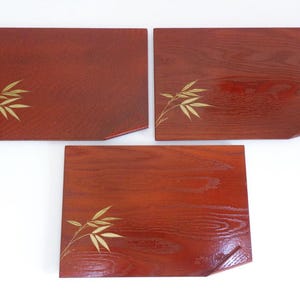 Puede incluir: Tres bandejas rectangulares de madera marrón rojizo con un acabado brillante. Cada bandeja presenta un diseño de hojas de bambú de color dorado en la esquina. Las bandejas tienen un borde angulado único en un lado, agregando un toque distintivo a su diseño.