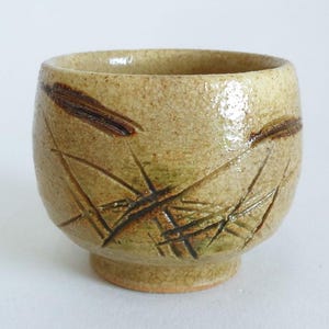 May include: A handmade ceramic cup with a beige and brown color scheme. The cup features a textured surface and is adorned with etched plant-like designs. The cup has a rounded shape and a small, circular base.