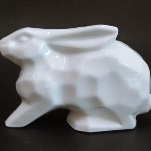 Op de afbeelding: Een witte keramische konijnenfiguur met een geometrisch ontwerp. Het konijn zit in een zittende positie met zijn oren omhoog. Het beeldje is glad en glanzend, met een minimalistische esthetiek.