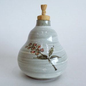Op de afbeelding: Een grijze keramische fles met een bloemenontwerp en een houten stop. De fles heeft een peervormig lichaam en een smalle hals.