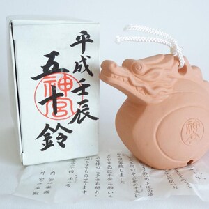 Puede incluir: Una campana de cerámica color terracota con forma de dragón y un cordón blanco. Al lado hay una caja de cartón blanca con caligrafía japonesa negra y un sello rojo. Debajo se encuentra un pequeño papel con texto en japonés.