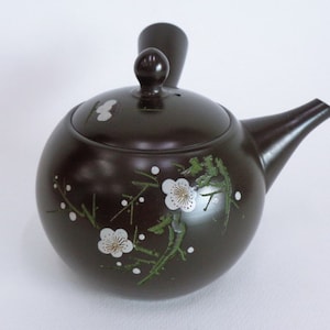 Könnte beinhalten: Eine dunkelbraune Teekanne aus Keramik mit Deckel und Ausguss. Die Teekanne ist mit weißen Blüten und grünen Blättern verziert.