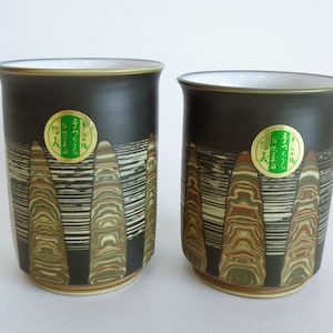 May include: Two black ceramic mugs with a striped pattern in shades of brown, green, and white. The mugs have a gold rim and a green and gold label with Japanese text.
