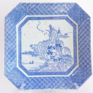 May include: An octagonal porcelain plate with a blue and white landscape design. The plate features a central scene of mountains, trees, and a small building, surrounded by a geometric border. The plate is supported by a clear plastic stand.
