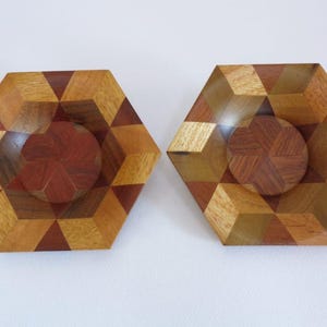 Puede incluir: Dos cuencos hexagonales de madera con patrones geométricos. Los cuencos presentan un diseño circular central y están hechos de varias tonalidades de madera, incluyendo marrones claros y oscuros. El diseño crea un efecto tridimensional.