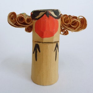 Könnte beinhalten: Eine Holzfigur mit rotem Gesicht, schwarzen Augen und schwarzen Augenbrauen. Die Figur hat zwei große, gekräuselte Holzstücke auf beiden Seiten des Kopfes, die an Haare erinnern. Die Figur steht auf einem Holzsockel mit zwei schwarzen Pfeilen, die darauf gemalt sind.