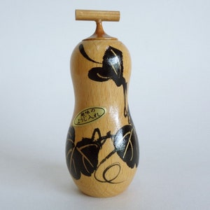 Puede incluir: Un tapón de botella de madera con un diseño de hojas negras y doradas. El tapón tiene una pequeña manija de madera en la parte superior y una etiqueta que dice "木の葉入り".