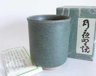 May include: A green ceramic tea cup with a textured surface, accompanied by a matching box and a small paper with Japanese text. The cup has a cylindrical shape with a slightly flared rim and a light-colored base.