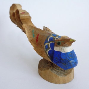 Könnte beinhalten: Eine hölzerne Vogelfigur mit einem blau-weiß bemalten Körper und einem langen, gekräuselten Schwanz. Der Vogel hat ein grünes Auge und einen gelben Schnabel.