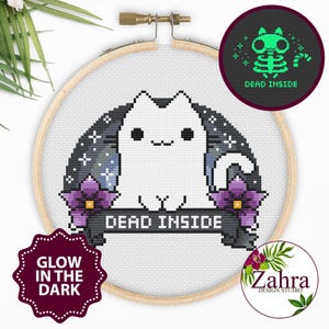 May include: A cross-stitch embroidery in a round wooden hoop featuring a white cat with black eyes and a mouth, flanked by purple flowers and a dark background with stars. A banner below the cat displays the text DEAD INSIDE in white letters.