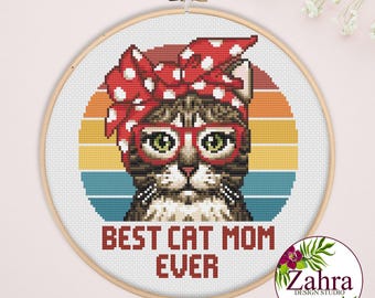May include: A cross-stitch pattern in a wooden hoop featuring a tabby cat wearing red glasses and a red polka-dot headscarf. The background is a sunset-colored circle with horizontal stripes. The text reads BEST CAT MOM EVER in red letters.