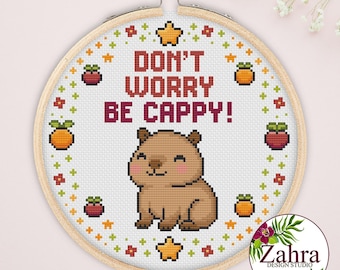 May include: A circular wooden embroidery hoop containing a cross-stitch pattern on white fabric. The design features a brown capybara, yellow stars, and small red and orange fruit icons, with the text DON'T WORRY BE CAPPY! in dark red letters.