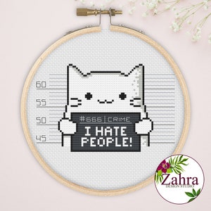Op de afbeelding: Kruissteekborduurwerk in een houten hoepel met een gepixeleerde witte kat die een bord vasthoudt met de tekst "I HATE PEOPLE!" en "#666 CRIME" erboven. De achtergrond heeft een hoogtediagram. Het logo van Zahra Design Studio staat rechtsonder.