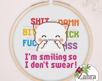 May include: A circular wooden embroidery hoop containing a cross-stitch design of a white cat with pink cheeks. Behind the cat are colorful swear words in various fonts. Below the cat, the text reads, I'm smiling so I don't swear! in pink.