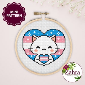 May include: A white cat with pink cheeks holding a transgender pride heart in its paws. The cat is surrounded by a blue and pink heart with white stars.