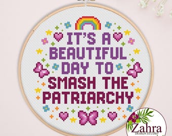 May include: A circular wooden embroidery hoop containing white fabric with a cross-stitch design. The text reads IT'S A BEAUTIFUL DAY TO SMASH THE PATRIARCHY in purple letters, surrounded by a colorful rainbow, pink butterflies, hearts, and stars.