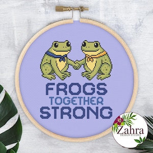 Op de afbeelding: Een borduurring met kruissteek met twee groene kikkers die elkaars hand vasthouden op een lichtpaarse achtergrond. De tekst "FROGS TOGETHER STRONG" is in blauw gestikt. De ring is van hout.