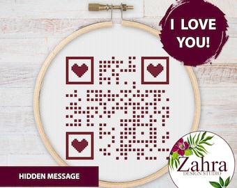 Puede incluir: Un bastidor de bordado circular de madera que contiene tela blanca con un patrón de código QR en punto de cruz de color rojo oscuro. El diseño presenta tres pequeños corazones de color rojo oscuro dentro de los cuadrados del código QR. El texto dice I LOVE YOU! y HIDDEN MESSAGE.