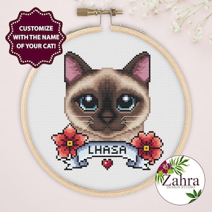 May include: A cross-stitch pattern in a wooden embroidery hoop featuring a Siamese cat with blue eyes, flanked by two red flowers. A light blue banner below the cat displays the name LHASA in black text, with a small red heart centered underneath.