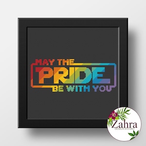 May include: A framed cross-stitch artwork with the text "MAY THE PRIDE BE WITH YOU" in rainbow colors. The artwork is set against a dark gray background and framed in a black square frame. A circular logo is in the bottom right corner.