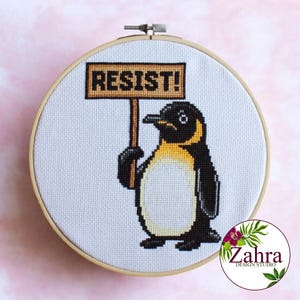 May include: A cross-stitch embroidery hoop with a white background and a cartoon penguin holding a sign that says "RESIST!" The penguin is black and white with a yellow beak and a yellow chest.