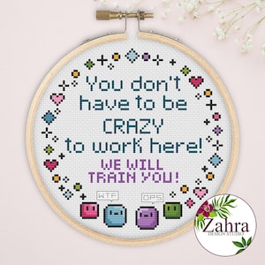 May include: A circular wooden embroidery hoop containing white fabric with cross-stitched text that reads: You don't have to be CRAZY to work here! WE WILL TRAIN YOU! Below the text are four small colourful square characters with speech bubbles.