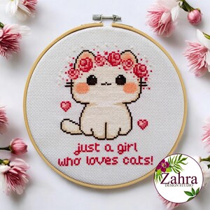 May include: A cross-stitch embroidery in a round wooden hoop featuring a white cat with pink cheeks wearing a crown of red roses. Two small red hearts flank the cat. The text below reads, "just a girl who loves cats!" in red lettering.