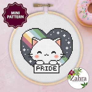 Pode incluir: Um mini padrão de ponto cruz com um gato branco dentro de um design em forma de coração. O coração é composto por um arco-íris e um padrão de noite estrelada. O gato segura uma placa que diz "PRIDE". O design está num bastidor de bordado de madeira.