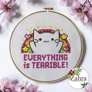 Könnte beinhalten: Ein runder Stickrahmen aus Holz mit einem Kreuzstichmotiv, das eine weiße Katze mit einem Regenbogen, gelben Sternen und violetten Herzen zeigt. Unter der Katze steht der Text EVERYTHING IS TERRIBLE! in fetten, dunkelrosa, pixeligen Buchstaben.
