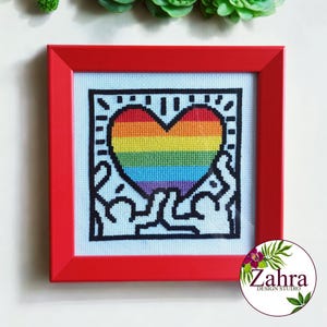 May include: A square cross-stitch artwork in a bright red frame featuring a central rainbow-colored heart. The heart is surrounded by black-outlined figures with raised arms on a white fabric background, creating a bold, graphic, and colorful display.