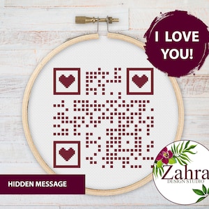 May include: A white embroidery hoop with a red and white QR code design. The QR code has three red hearts in the corners. The text "I LOVE YOU!" is written in a red circle above the hoop. The text "HIDDEN MESSAGE" is written below the hoop.