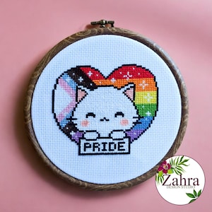 May include: A white cat with a rainbow heart around it holding a sign that says "PRIDE". The heart is made up of the colors of the rainbow flag, including black, brown, light blue, pink, and white.